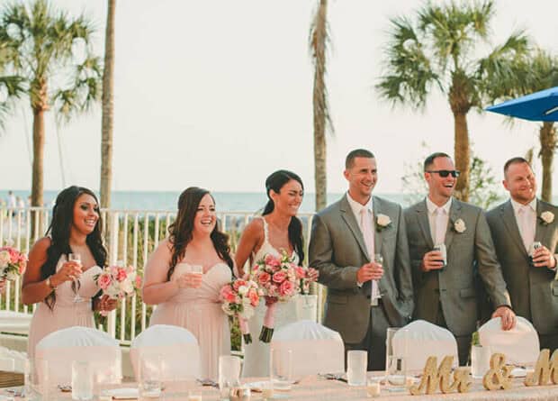 A wedding party stands behind a decorated outdoor table, holding drinks and bouquets, with palm trees and a fence visible in the background.