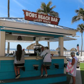 Outdoor beach bar with a turquoise counter, three people sitting and standing, palm trees, and lounge chairs in the background under a clear blue sky.