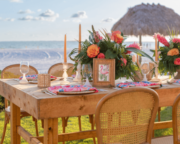 A wooden table set for a meal with floral centerpieces, candles, and tropical-themed napkins, situated outdoors near a beach with a thatched umbrella in the background.