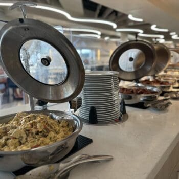 A buffet setup with chafing dishes containing food, stacked plates, and serving utensils on a marble counter in a brightly lit dining area.