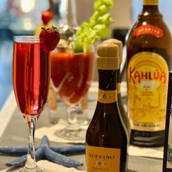 A glass of red sparkling cocktail with a strawberry garnish, a small bottle of Ruffino Prosecco, a bottle of Kahlúa, and a Bloody Mary with celery on a table.