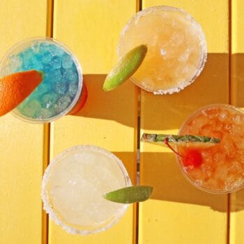 Four colorful cocktails with ice and fruit garnishes sit on a bright yellow table, casting shadows in sunlight.