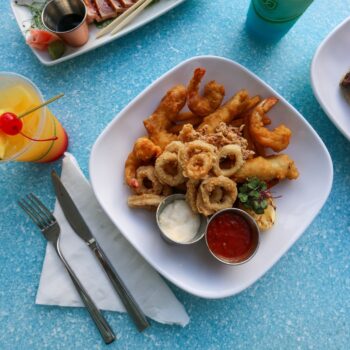 A white plate with fried shrimp, calamari, dipping sauces, lemon, and garnish on a blue table, next to a drink with a cherry and silverware on a napkin.