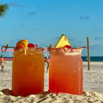 Two colorful cocktails in mason jars sit on sandy beach with ocean, palm tree, and people playing volleyball in background.