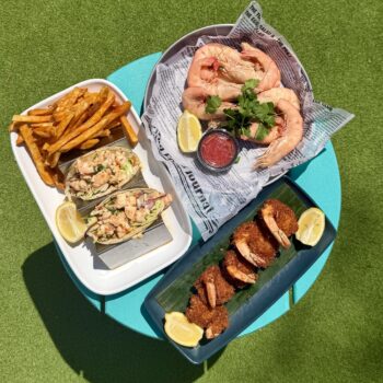 Three dishes on a round blue table: a plate of shrimp with cocktail sauce and lemon, shrimp tacos with fries, and breaded shrimp with lemon. The table is set on green grass.