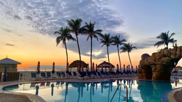 A poolside scene at sunset with palm trees, lounge chairs, umbrellas, a rock feature, and a view of the ocean in the background.