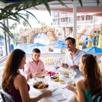 A family of four sits at a restaurant table by a window, eating and talking, with a pool and resort visible outside.