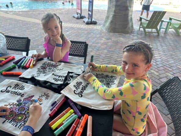 Two young girls sit at a table by a pool, coloring designs on 