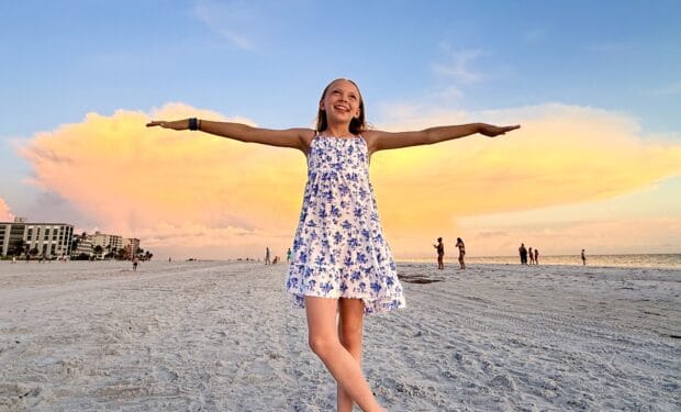 A girl in a white and blue dress stands on a sandy beach with arms outstretched, a large cloud and sunset sky in the background. People and buildings are visible in the distance.