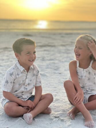 Two young children sit barefoot on a sandy beach at sunset, smiling and laughing together.