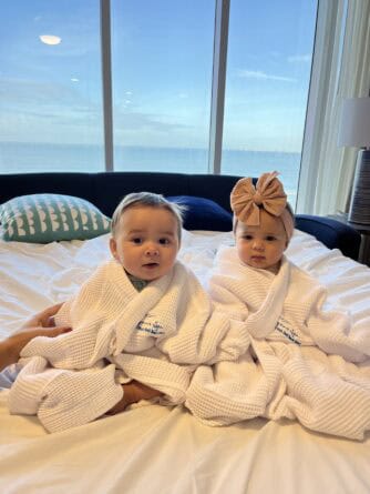 Two babies in white bathrobes sit on a bed by a large window with an ocean view; one wears a tan headband with a bow.