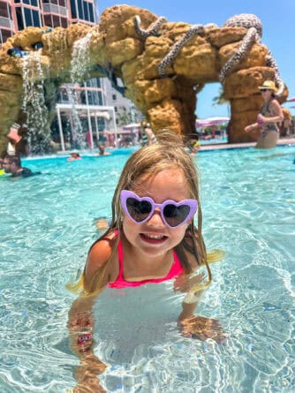 A young girl in heart-shaped sunglasses and a pink swimsuit smiles while playing in a shallow pool; artificial rock formations and waterfalls are in the background.