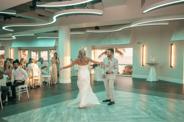 A bride and groom dance together in a modern indoor venue with guests seated around them and large windows showing a sunset over palm trees.