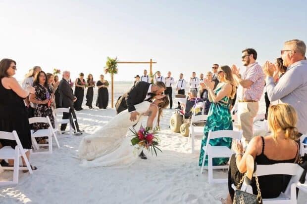 A bride and groom kiss at the end of the aisle on a sandy beach, surrounded by applauding wedding guests seated on white chairs.