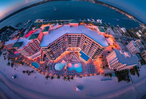 Aerial view of a large beachfront resort with pools and palm trees, illuminated at dusk, facing a calm body of water with boats and a sandy shoreline.