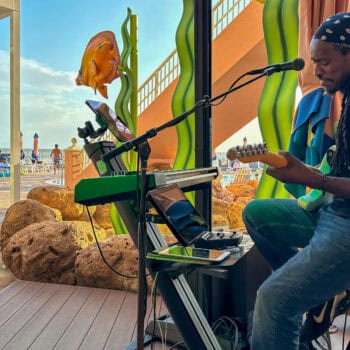 A musician with a guitar and keyboard performs indoors near large aquarium decor in a poolside area, with people and tables visible outside.