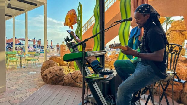 A musician with a guitar and keyboard performs indoors near large aquarium decor in a poolside area, with people and tables visible outside.