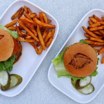 Two plates each with a burger, seasoned fries, and sliced pickles on a white surface; burgers have lettuce and a grilled logo on the bun.