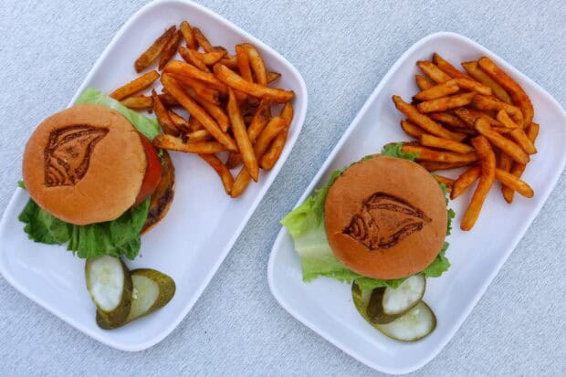 Two plates each with a burger, seasoned fries, and sliced pickles on a white surface; burgers have lettuce and a grilled logo on the bun.