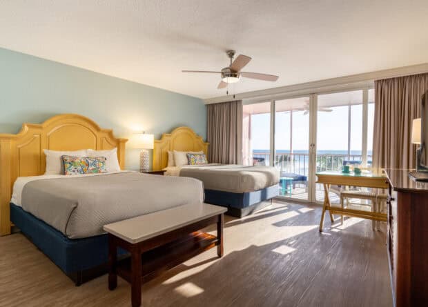 Hotel room with two double beds, a ceiling fan, a bench, a TV, and a sliding glass door leading to a balcony with an ocean view.