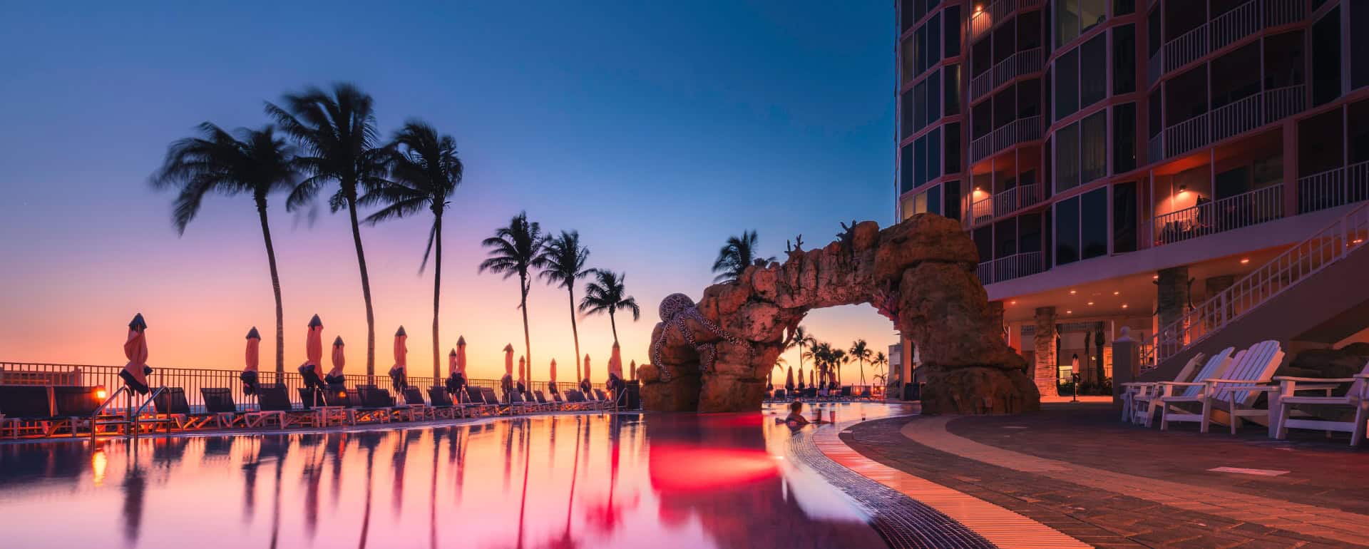 Hotel pool at sunset with palm trees, lounge chairs, and a rock arch feature; lights reflect on the calm water near a modern building.