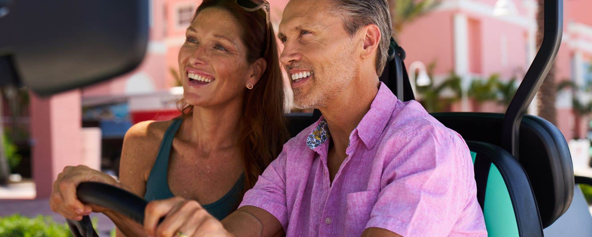 A smiling man and woman ride together in a golf cart on a sunny day, with pink buildings and palm trees in the background.