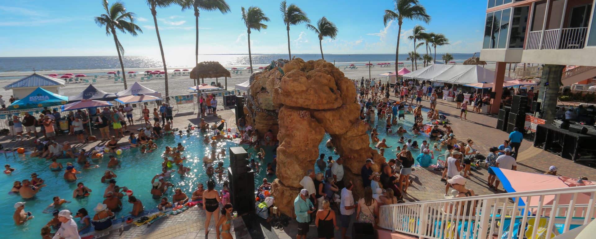 Crowd of people gathered around a pool near the beach, with palm trees, tents, and a rocky structure, under a sunny sky.