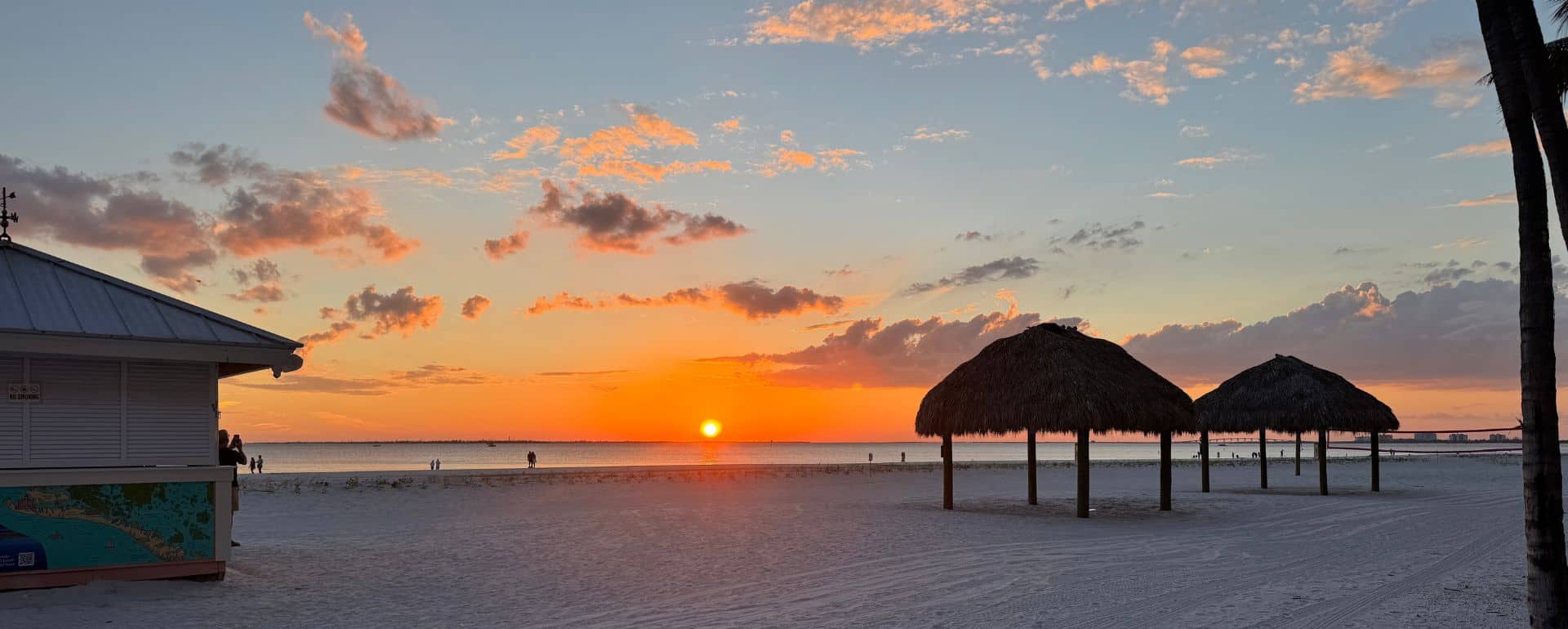 Sunset over a sandy beach with a closed kiosk on the left and two thatched-roof shelters on the right; a few clouds are scattered in the sky.