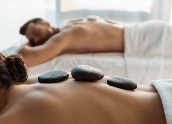 Two people lying face down on massage tables with towels, each with hot stones placed on their bare backs in a spa setting.