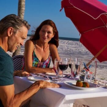 A man and woman sit at an outdoor table on a sandy beach, dining under a pink umbrella with food, wine glasses, and silverware on the table.