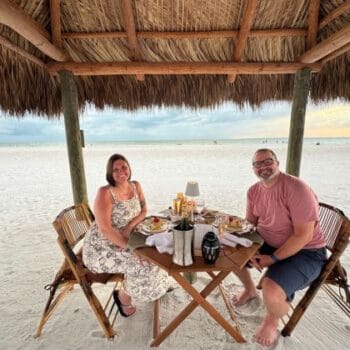 Two people sit at a wooden table under a thatched-roof gazebo on a sandy beach, enjoying a meal with drinks as the sun sets in the background.