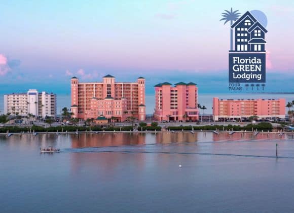 Waterfront view of pink and beige hotels at sunset with a "Florida Green Lodging Four Palms" logo in the sky corner, and a boat moving on the water in the foreground.