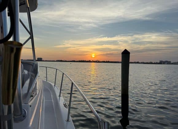 A boat is docked on calm water at sunset, with a single piling in the foreground and buildings visible on the distant shoreline under a partly cloudy sky.
