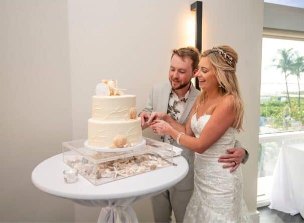 A bride and groom in wedding attire cut a two-tiered cake decorated with seashells and starfish at a reception with a beach view.