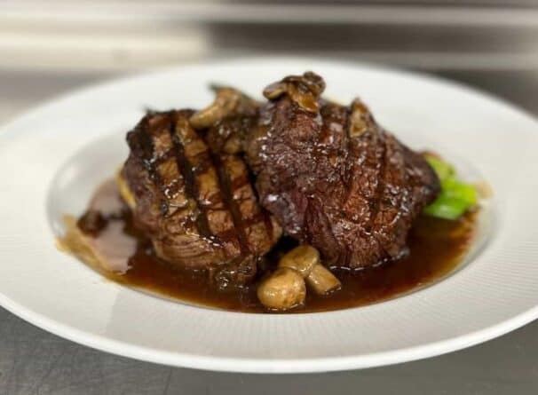 Two grilled steak medallions topped with mushrooms and brown sauce, served on a white plate.