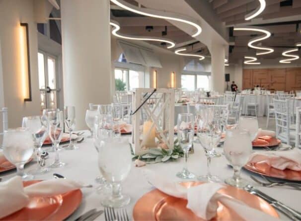 A decorated banquet hall with round tables set with pink plates, white napkins, glassware, and a lantern centerpiece, under modern ceiling lights.