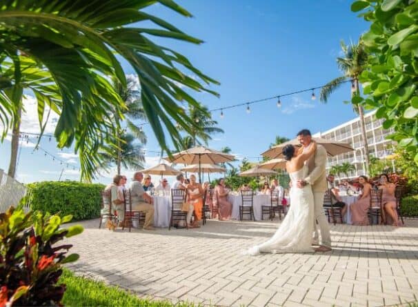 A bride and groom share their first dance outdoors at a sunny wedding reception, surrounded by seated guests and tropical greenery.