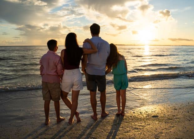 Four people stand barefoot on the beach, facing the ocean and watching the sunset, with their backs to the camera.
