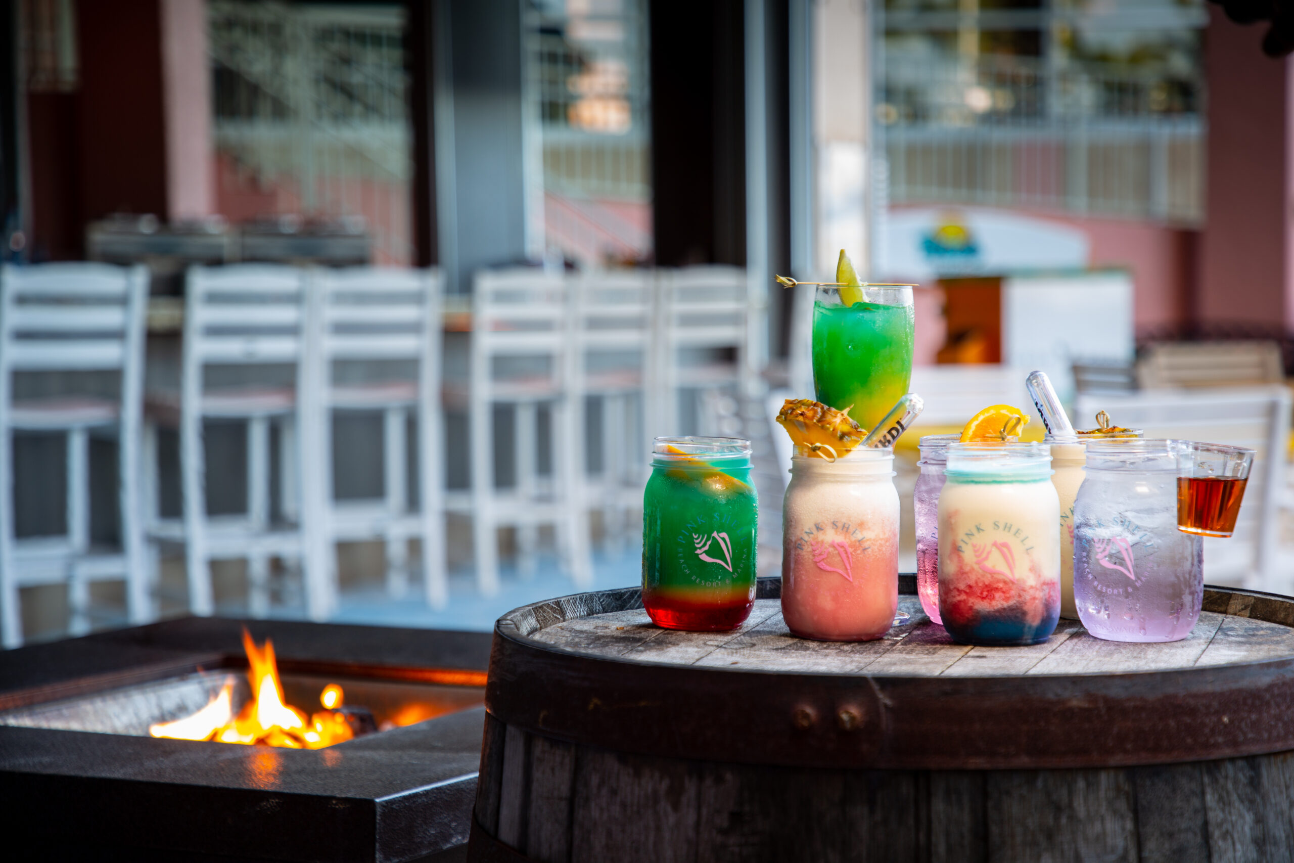 Five colorful cocktails in mason jars and a glass are arranged on a wooden barrel beside a fire pit, with empty chairs and tables in the background.