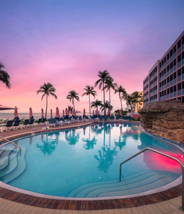 Outdoor hotel pool with curved edges, surrounded by lounge chairs and palm trees, next to a multi-story building at sunset with a pink and purple sky.