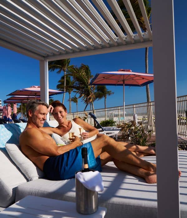 A man and woman relax on a shaded lounge chair by a pool, smiling and holding drinks, with umbrellas and palm trees in the background.