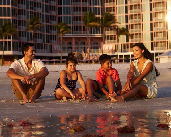A family of four sits on the beach, smiling and talking, with high-rise buildings and palm trees in the background.