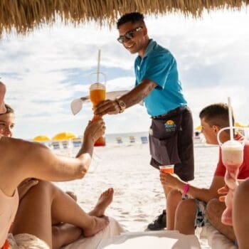 A beach server hands a tropical drink to a woman relaxing with friends under a thatched umbrella; yellow umbrellas and white sand are in the background.
