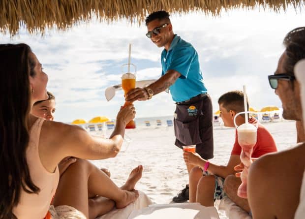 A beach server hands a tropical drink to a woman relaxing with friends under a thatched umbrella; yellow umbrellas and white sand are in the background.