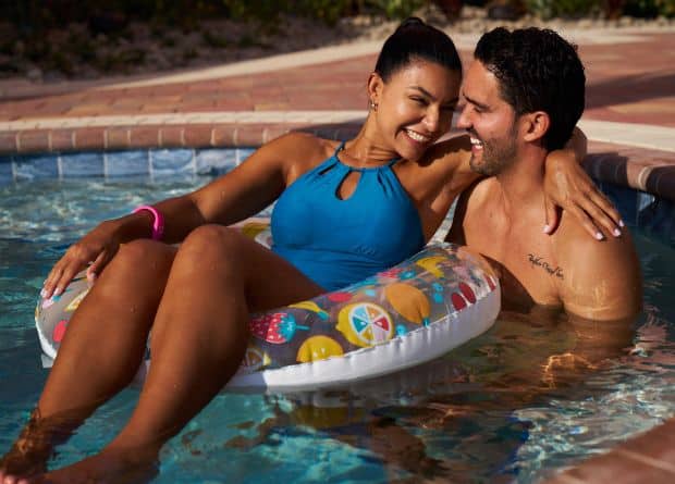 A woman in a blue swimsuit sits on a pool float, smiling at a man next to her in the pool. Both appear relaxed and happy, with sunlight illuminating the scene.