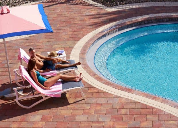 Two people are sunbathing on lounge chairs by a pool, under a pink umbrella, with towels, a hat, sunglasses, and a book nearby.