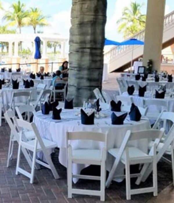 Round tables with white tablecloths and black napkins are set up with white chairs outdoors, near a palm tree, under a covered area.