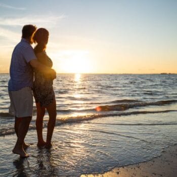 A couple stands barefoot on a beach, facing the ocean and watching the sunset together.
