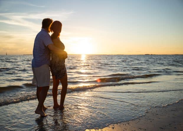 A couple stands barefoot on a beach, facing the ocean and watching the sunset together.