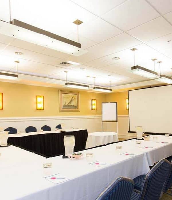 Conference room with U-shaped table arrangement, blue chairs, pitchers of water, notepads, a projector screen, and a whiteboard under ceiling lights.
